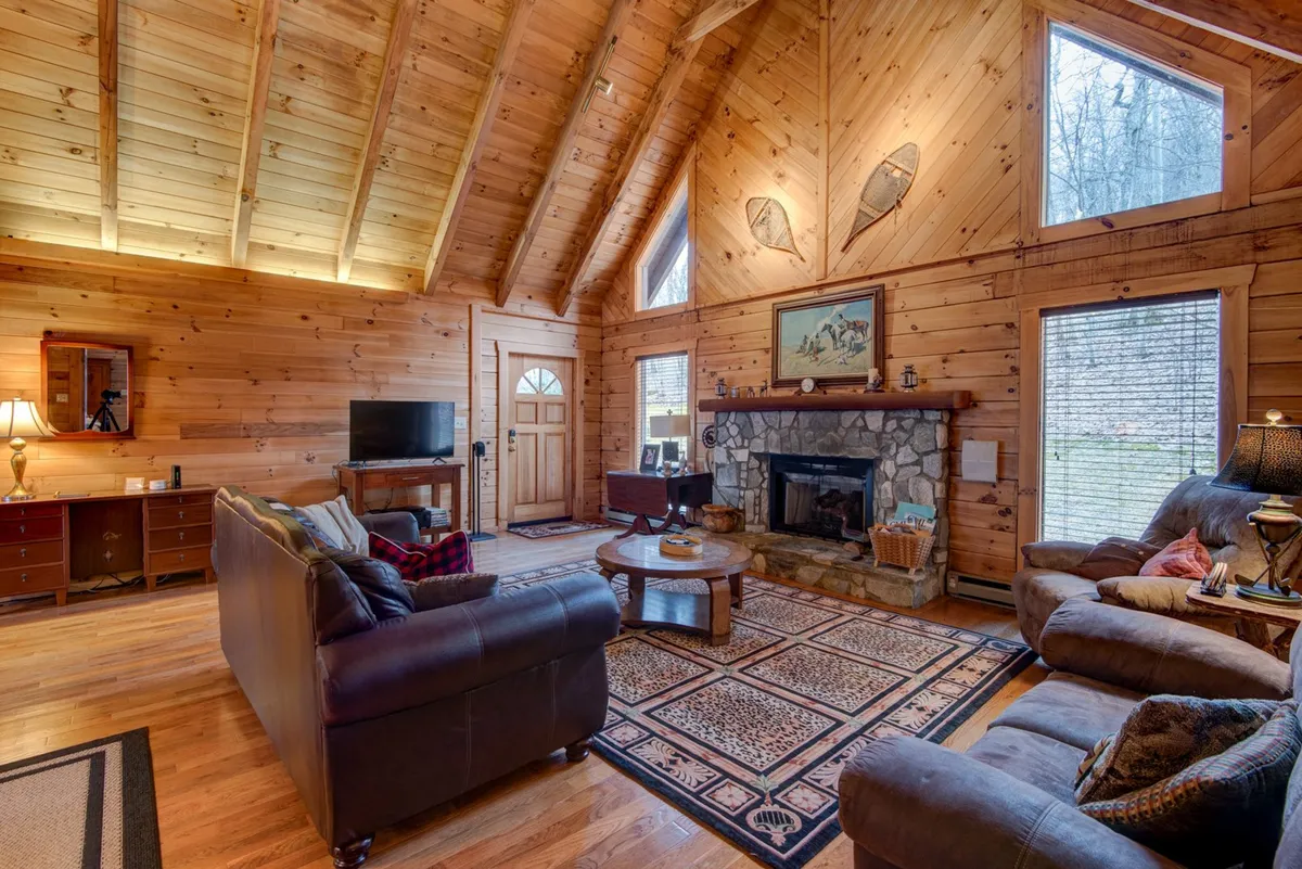 A welcoming living room featuring a stone fireplace, large windows that let in ample natural light, wooden beams, and comfortable sofas gathered around a cozy rug. The space feels inviting and is visually connected to the outdoors through the expansive windows, suggesting a connection to the fresh mountain air mentioned in the section.