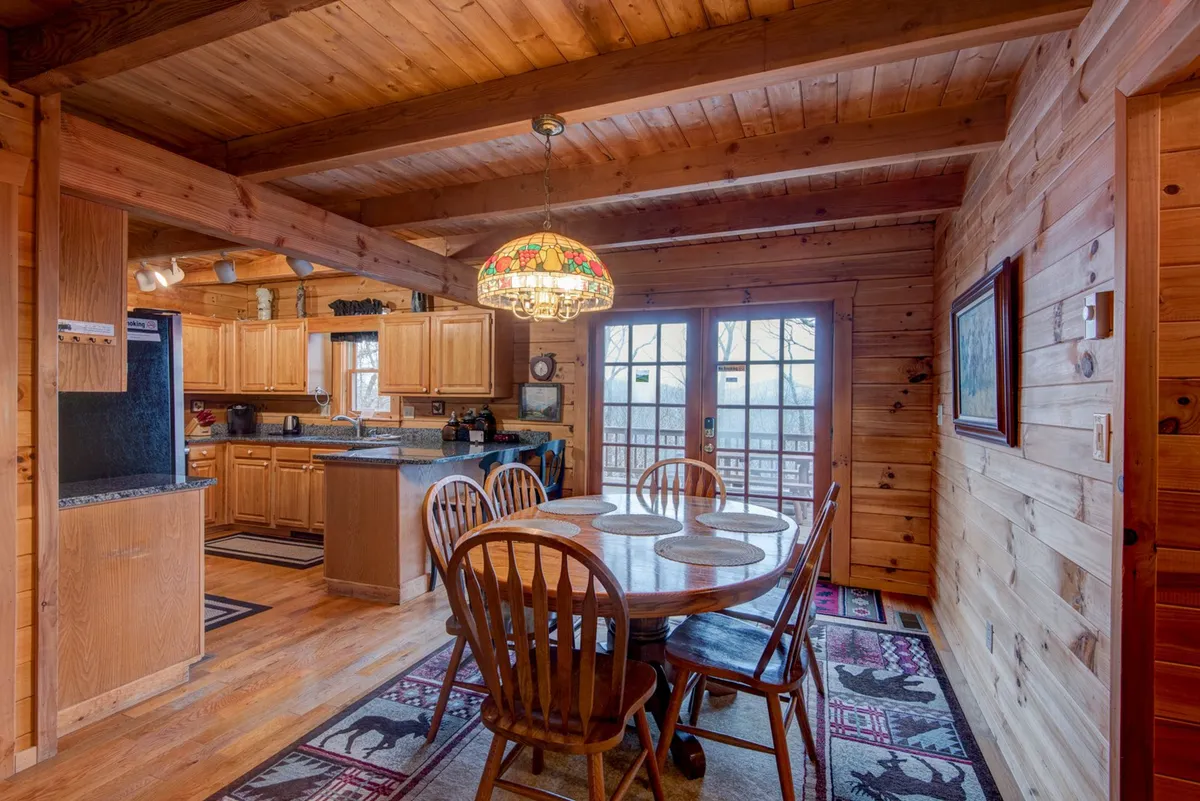 An inviting dining area with a wooden dining table beneath a Tiffany-style stained glass chandelier. In the background, you can spot the kitchen’s granite countertops and cabinetry, as well as French doors that open to the deck and let in ample natural light.