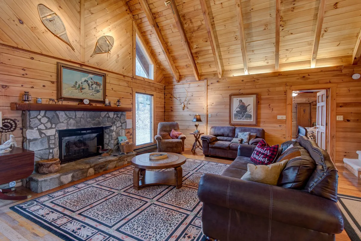 A stunning great room featuring a dramatic stone fireplace, cathedral ceilings with exposed wooden beams, and a plush, cozy seating area arranged for conversation or relaxation. Decorative artwork adds character while soft lighting enhances the room’s inviting warmth, perfect for unwinding or gathering with loved ones.
