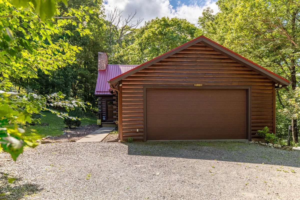 A welcoming log cabin with a warm red roof nestled amid lush greenery. The cabin's inviting porch and natural surroundings hint at peaceful mornings and private outdoor moments, while its sturdy structure suggests coziness inside. The garage door and ample space around the cabin suggest easy arrival and direct access to nature.