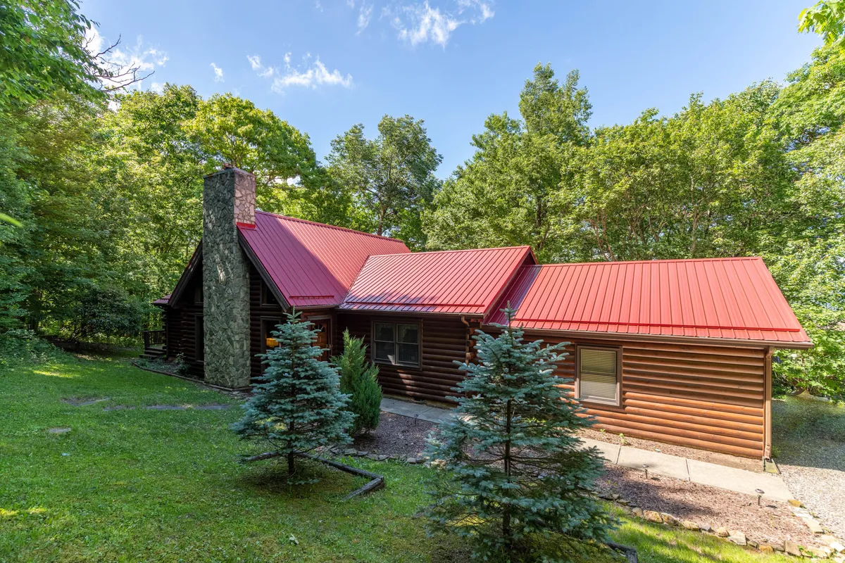 The hero image showcases a classic log cabin with a striking red metal roof and a stone chimney, nestled among lush trees and a beautifully landscaped yard. The setting is peaceful and inviting, with plenty of space for outdoor relaxation and gatherings, suggesting opportunities for stargazing and enjoying nature.