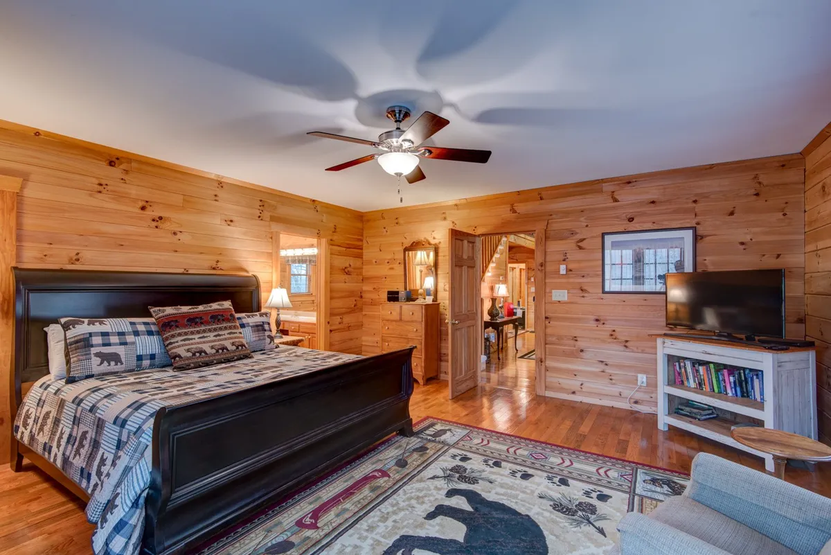 A bright, inviting sleeping area with a bed adorned in a patterned blanket, rustic wooden walls, and a cozy rug featuring a bear motif. The setting is enhanced by a ceiling fan, creating an atmosphere ideal for relaxing, reading, or watching TV.