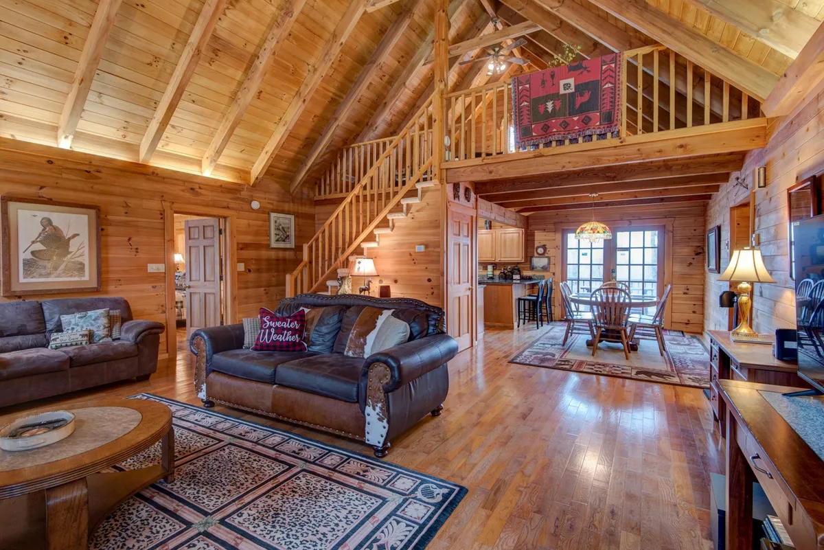 A wide view of the great room showcasing exposed wooden beams, a comfy sofa, an adjacent dining area, and a staircase. The space is styled for both cozy winter lounging and summer entertaining, perfect for family movie nights or group games.