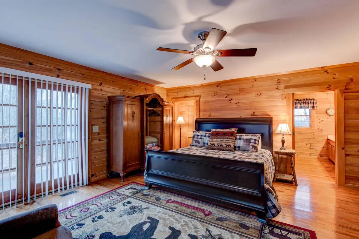 A serene bedroom featuring a large, inviting bed covered in soft linens, with a cozy rug at its base. Large windows let in natural light and offer views of the surrounding landscape. A wardrobe stands to one side, providing ample storage, and the overall atmosphere is peaceful and private.