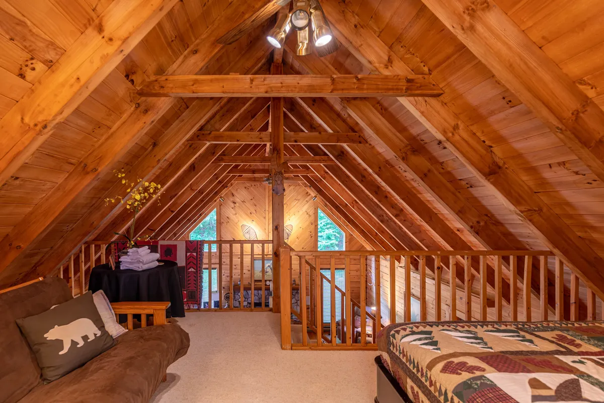 A warm loft space featuring wooden beams, a cozy seating area, and a bed with a quilt. The window allows for stargazing or enjoying natural light, and the seating area creates a welcoming nook for relaxation, reading, or movie nights.