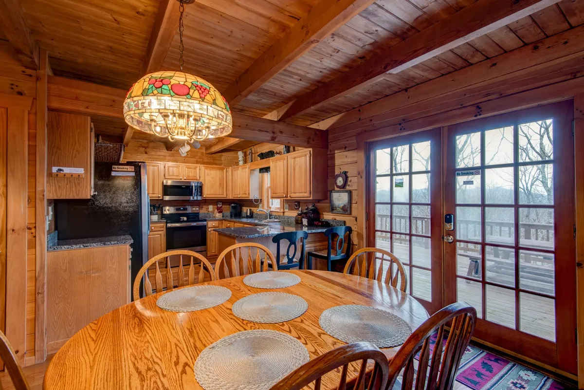 A warm, inviting dining area featuring a large wooden table surrounded by chairs, a colorful chandelier overhead, and double doors opening out to the deck. The well-equipped kitchen cabinetry is visible, suggesting ease for family meals and gatherings.