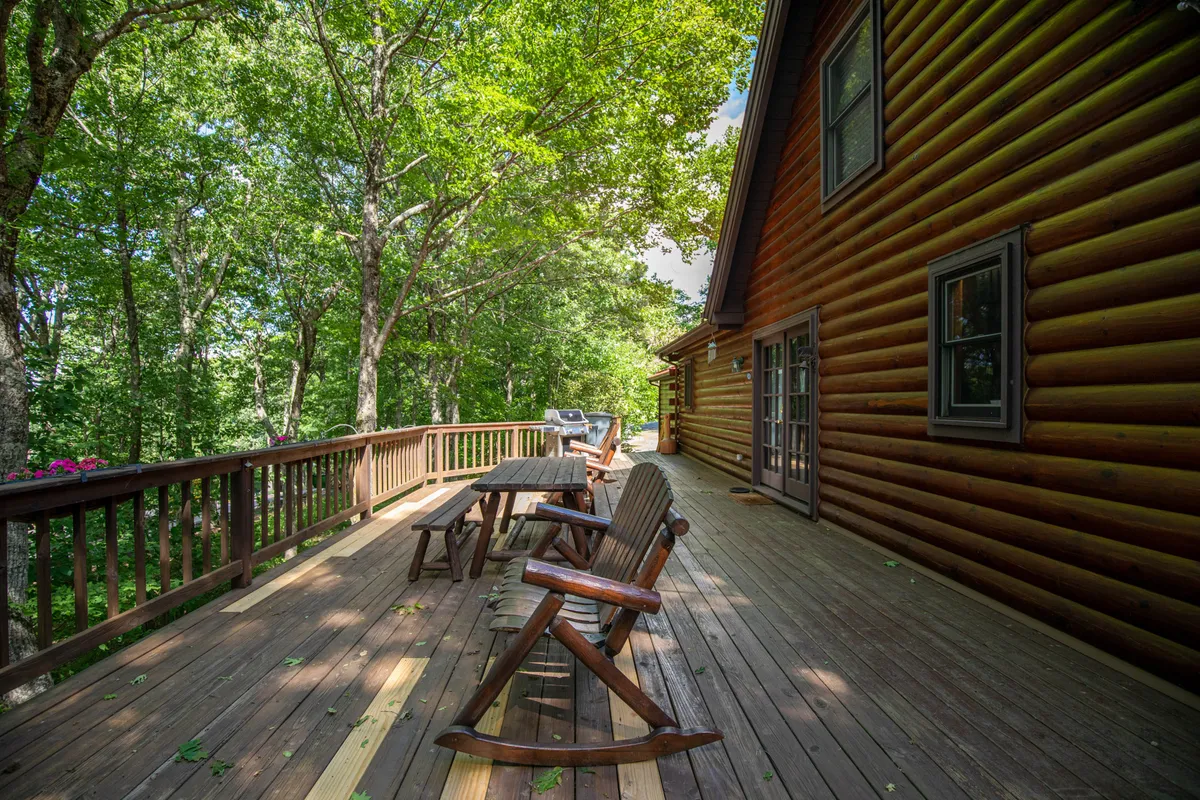 This image shows a spacious wooden deck attached to a log cabin, nestled among mature trees. The deck features outdoor seating and ample space for gatherings, with the tranquil beauty of the surrounding High Country landscape visible. The atmosphere is serene and inviting, emphasizing relaxation and connection with nature.